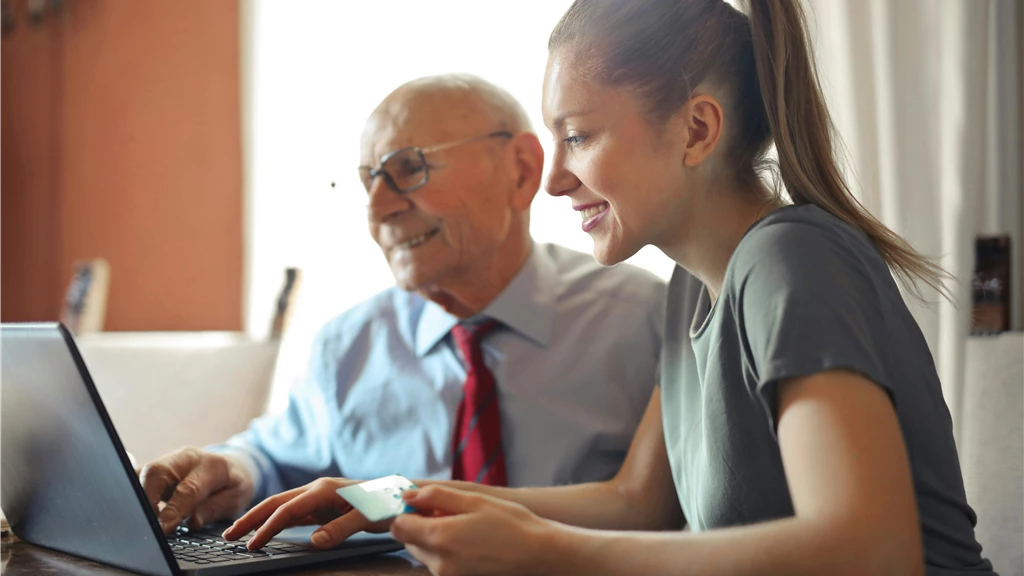 Young woman in casual clothes helping senior man in formal shirt with paying credit card in Internet using laptop while sitting at table