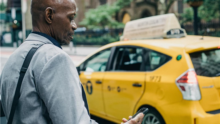 Businessman checks his phone while standing next to a yellow New York taxi on a city street.